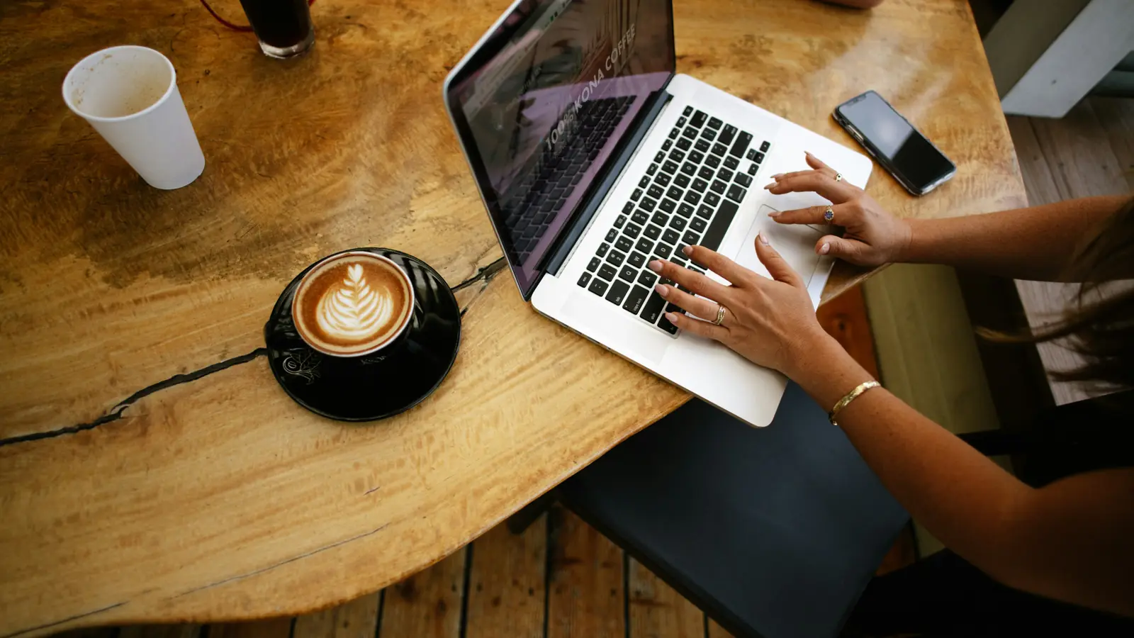Hands typing on a laptop at a café table, next to a coffee and a phone